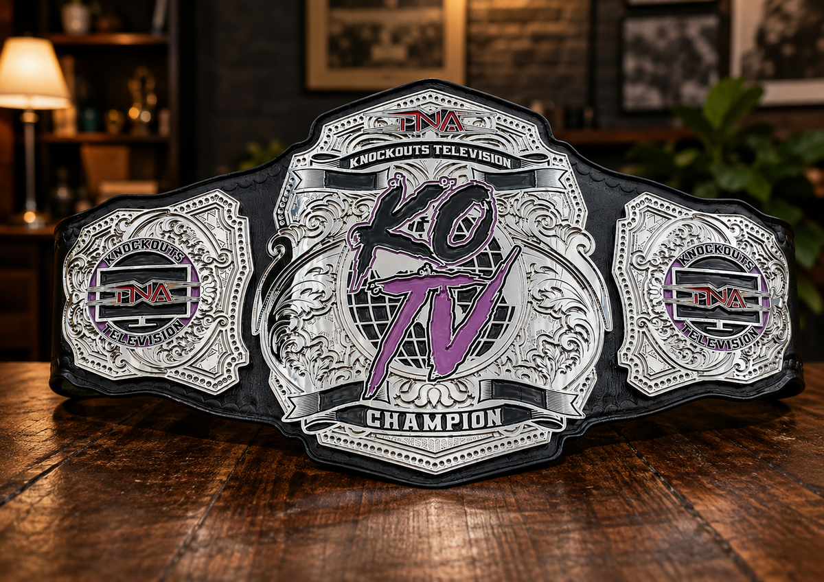 A large ornate championship belt displayed on a wooden table in a dimly lit indoor setting. The belt has a black base with silver-toned engraved plates and purple and red logo details in the center and on the side panels.