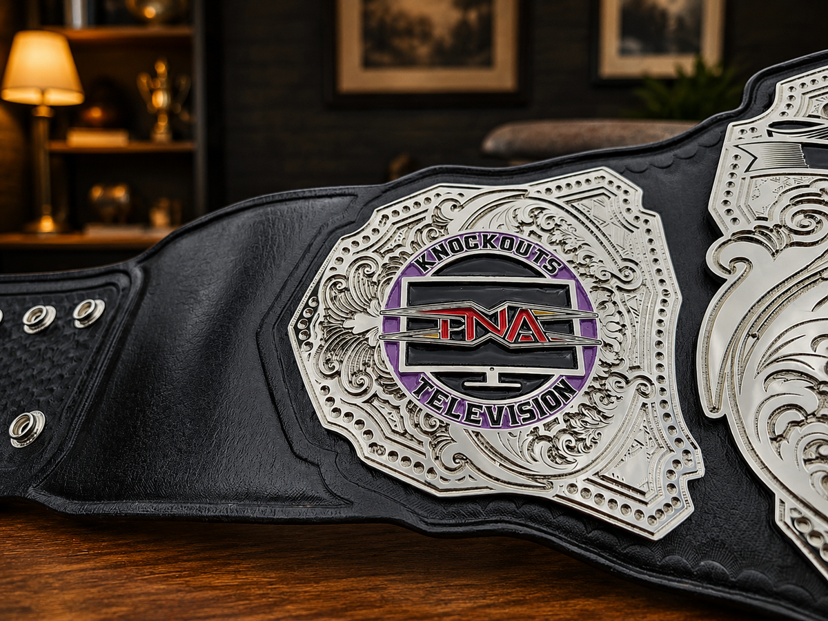 A close-up of a black leather championship belt with ornate silver metal plates, featuring a central circular badge with the text 'Knockouts Television' and 'PNA' in red and purple.