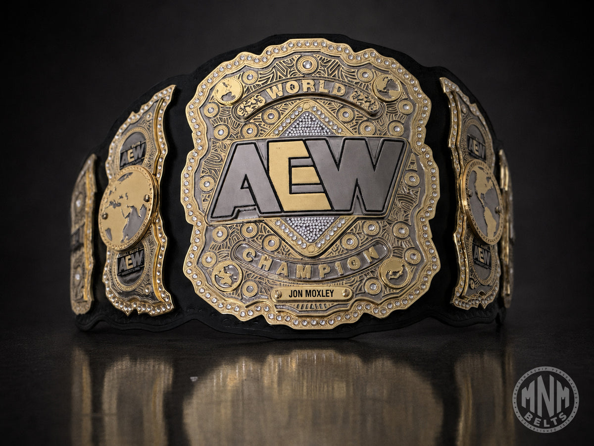 An AEW World Champion belt featuring ornate gold and silver plates on a black strap, displayed on a dark reflective surface with a watermark in the corner.