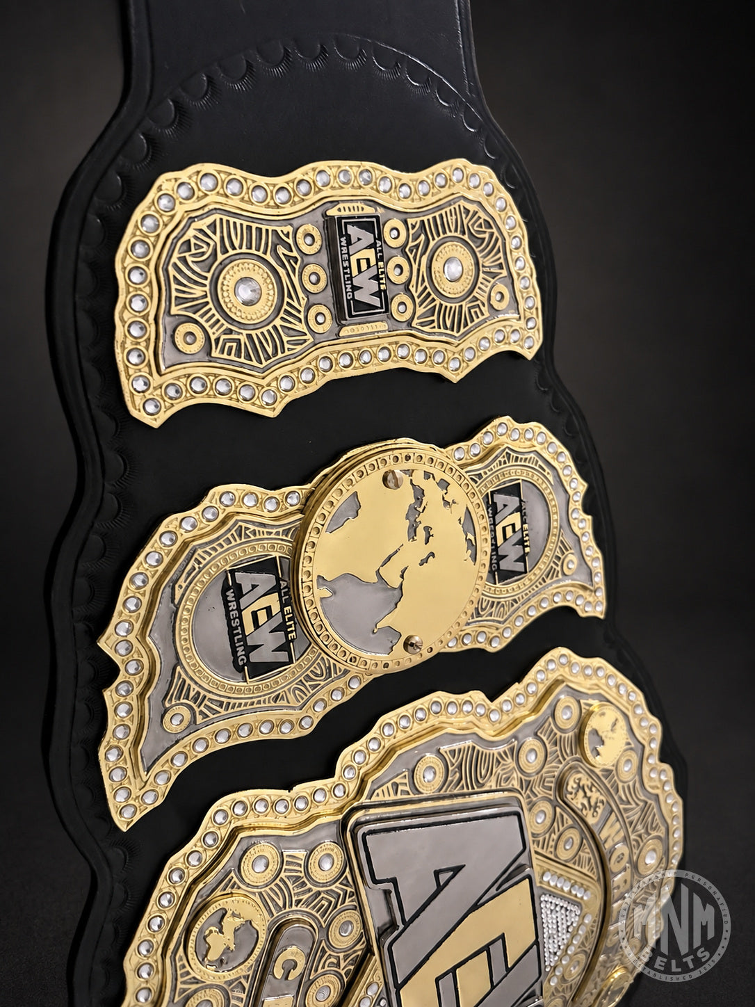 A close-up of a black and gold AEW (All In The Night) championship belt with ornate gold plates, silver details, and rhinestone accents on a dark background.