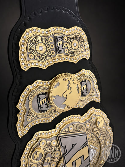 A close-up of a black and gold AEW (All In The Night) championship belt with ornate gold plates, silver details, and rhinestone accents on a dark background.