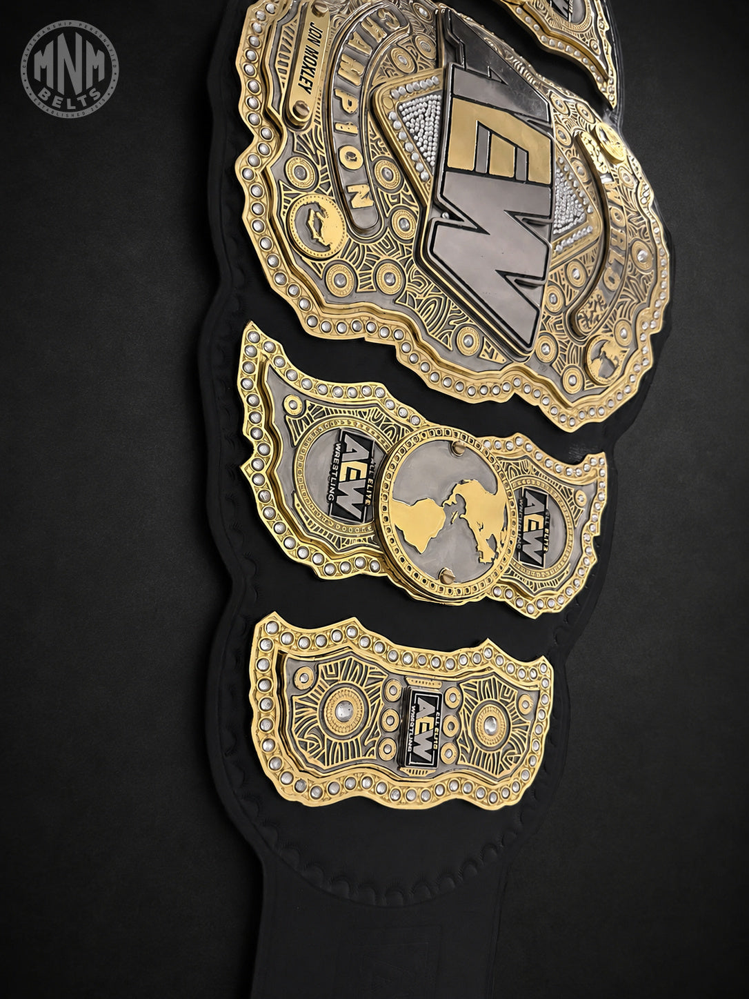 A close-up of a black championship belt with ornate gold and silver plates featuring the AEW logo and intricate detailing, set against a dark background.