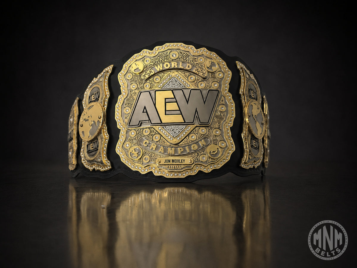 A gold and black AEW World Champion belt displayed on a dark reflective surface, with the central plate featuring the AEW logo and the name Jon Moxley.